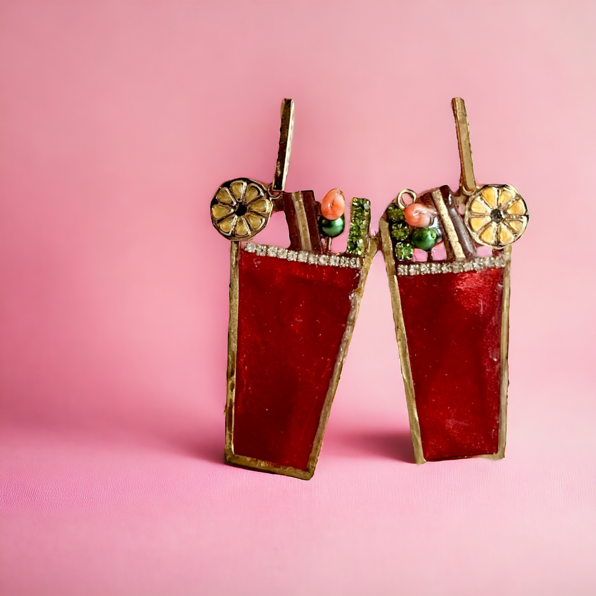 statement cocktail earrings by brand gumptious this pair of bloody mary earrings in a studio setting with a pink background are with a brass bezel and red resin, complete with a tiny resin and brass piece of bacon, a lemon wheel, a tiny skewer of shrimp and an olive complete with a celery stick and hang from a brass straw-like push back finding